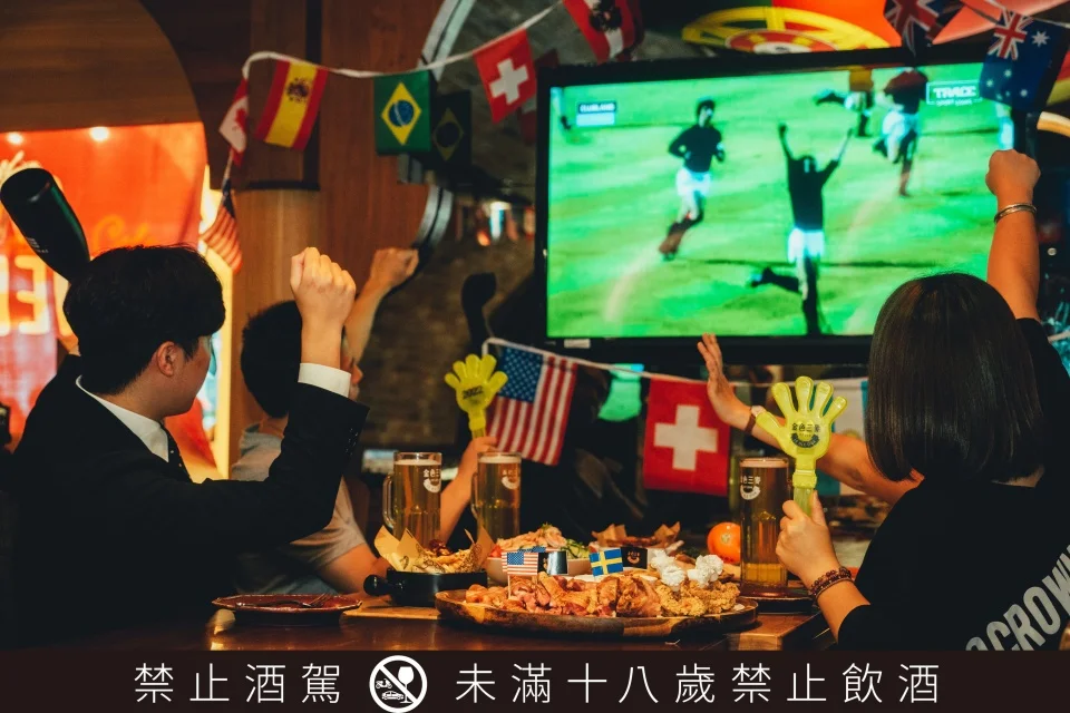 球迷在掛滿各國國旗的運動酒吧看世足，畫面下方標示禁止酒駕與未滿十八歲禁止飲酒